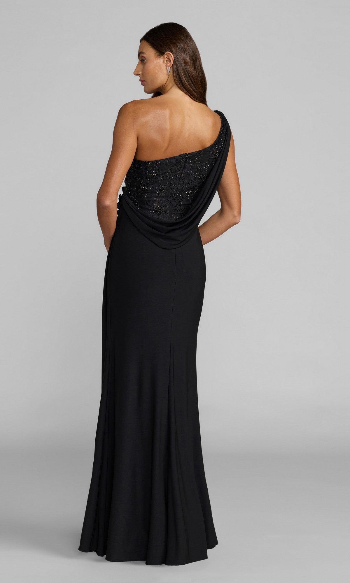 A woman stands with her back to the camera, wearing the Mac Duggal 13210 long formal dress—a floor-length, one-shoulder black gown featuring beaded detailing on the bodice and elegant 3D flower accents.